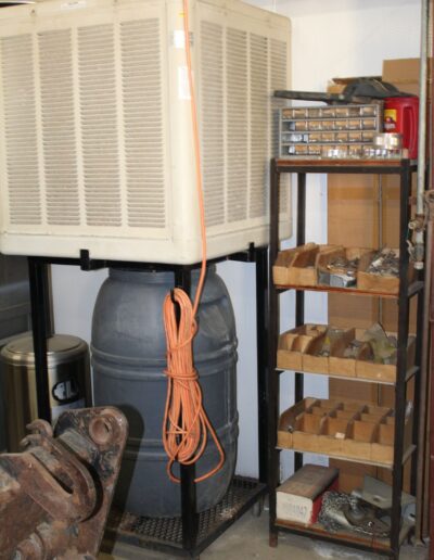 An orange extension cord lies on a cart with a large grey plastic barrel and a Port-A-Cool swamp cooler. Underneath is a shelf containing boxes and various parts.